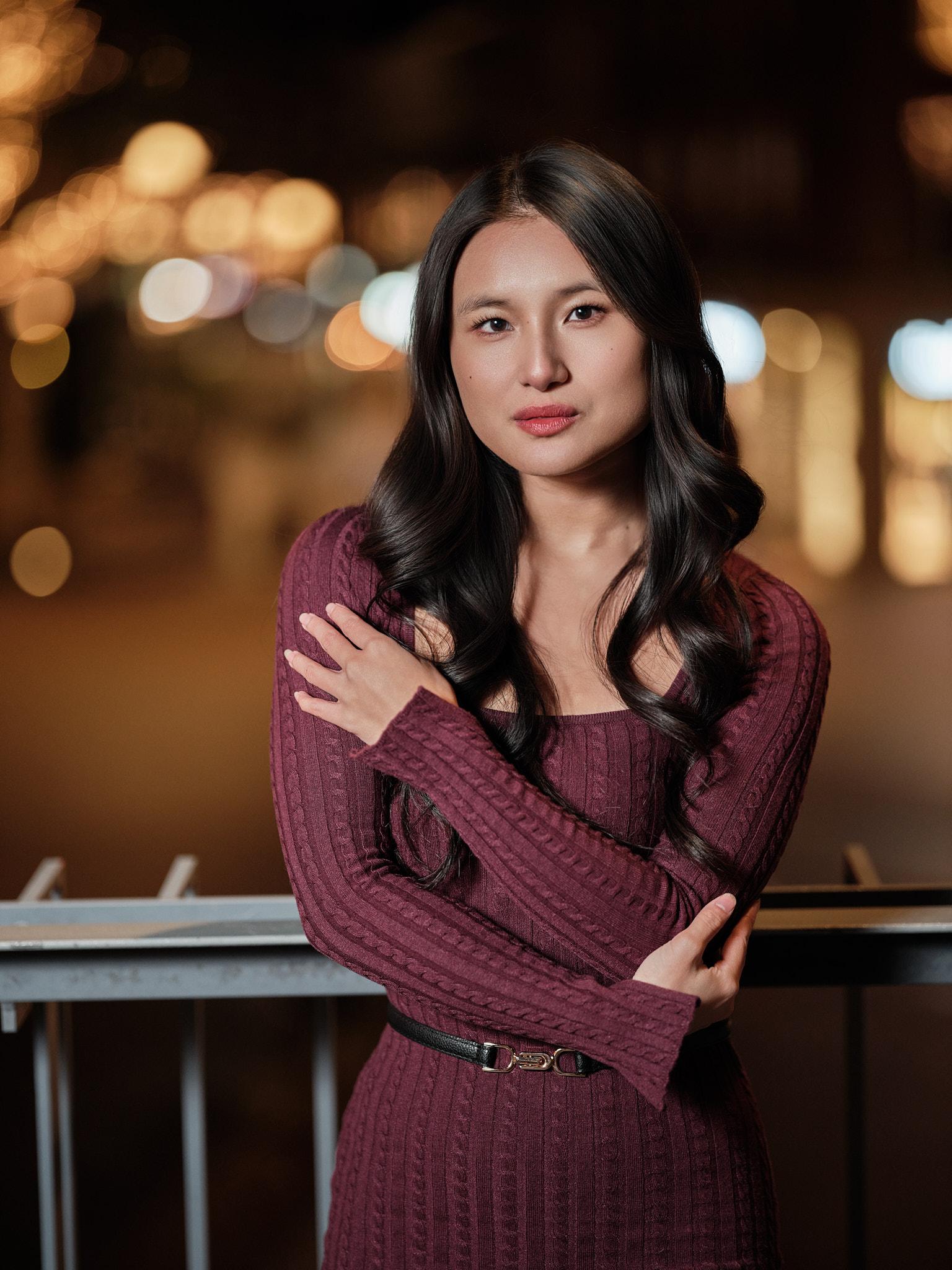 Intiem avondportret met zachte bokehlichten Avondportret van een jonge vrouw in een bordeauxrode jurk met armen over elkaar, staand tegen een railing met zachte stadslichten op de achtergrond.