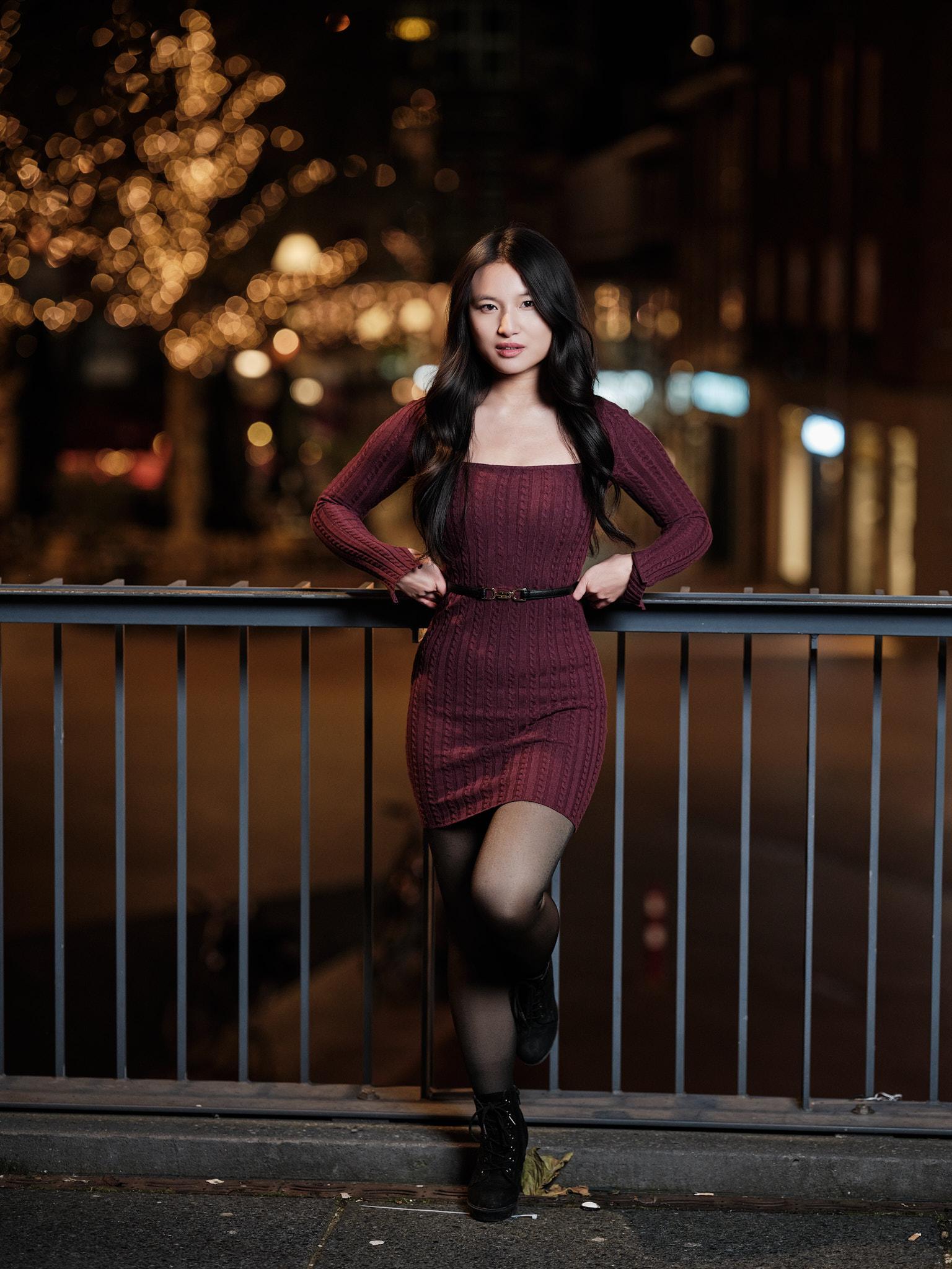 Fashion-achtig avondportret op straatlocatie Volledig avondportret van een jonge vrouw in bordeauxrode jurk, leunend tegen een railing met verlichte bomen en stadslichten op de achtergrond.